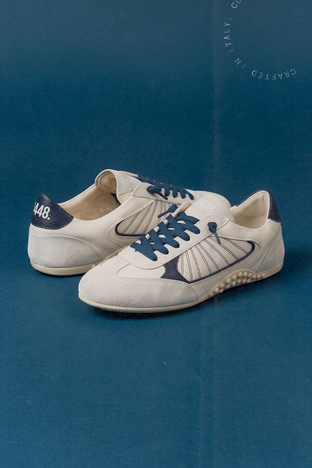 P448 low-top retro runner-style sneakers in cream leather and grey suede with navy blue laces and details on a cobalt blue background.