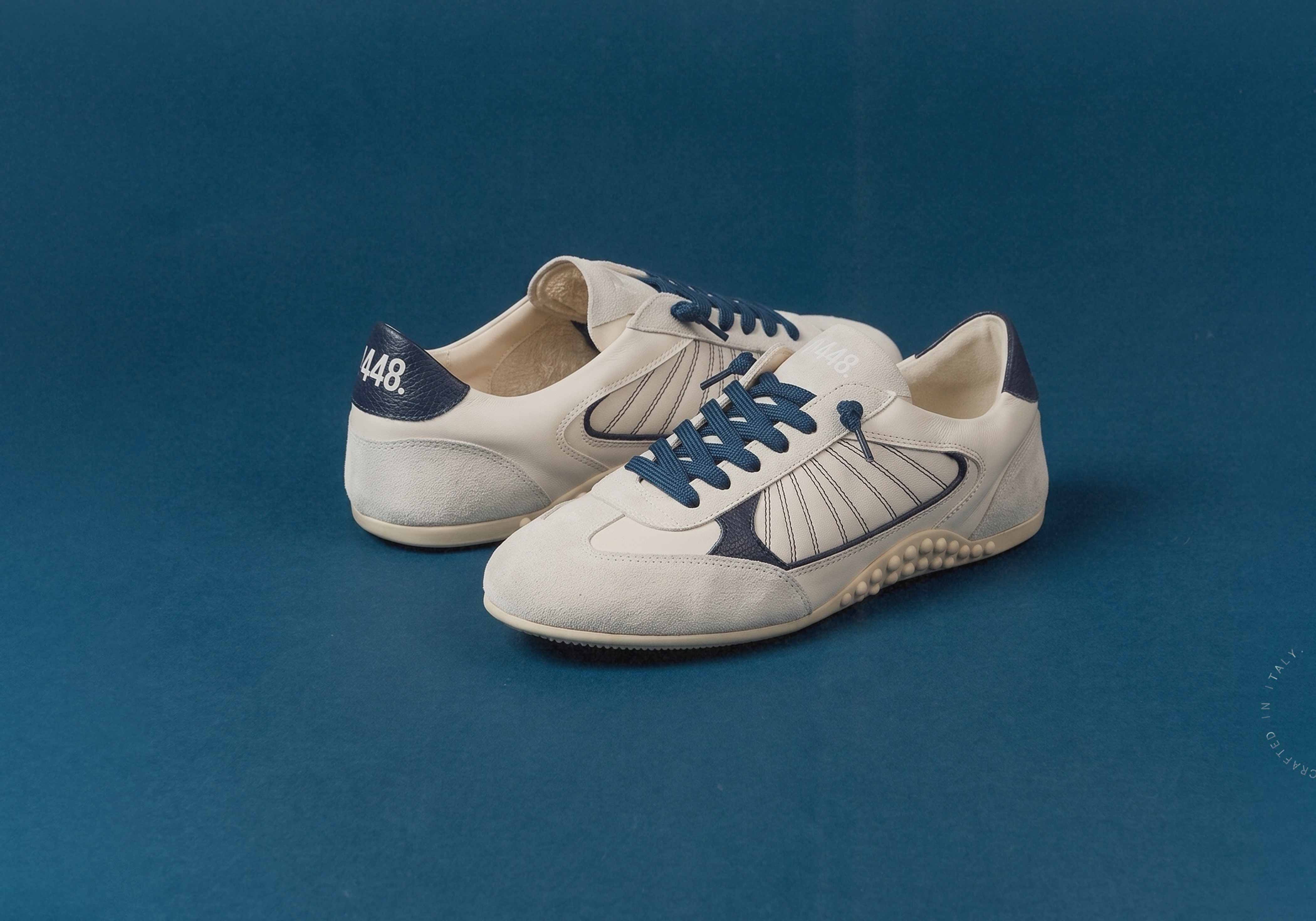 P448 low-top retro runner-style sneakers in cream leather and grey suede with navy blue laces and details on a cobalt blue background.