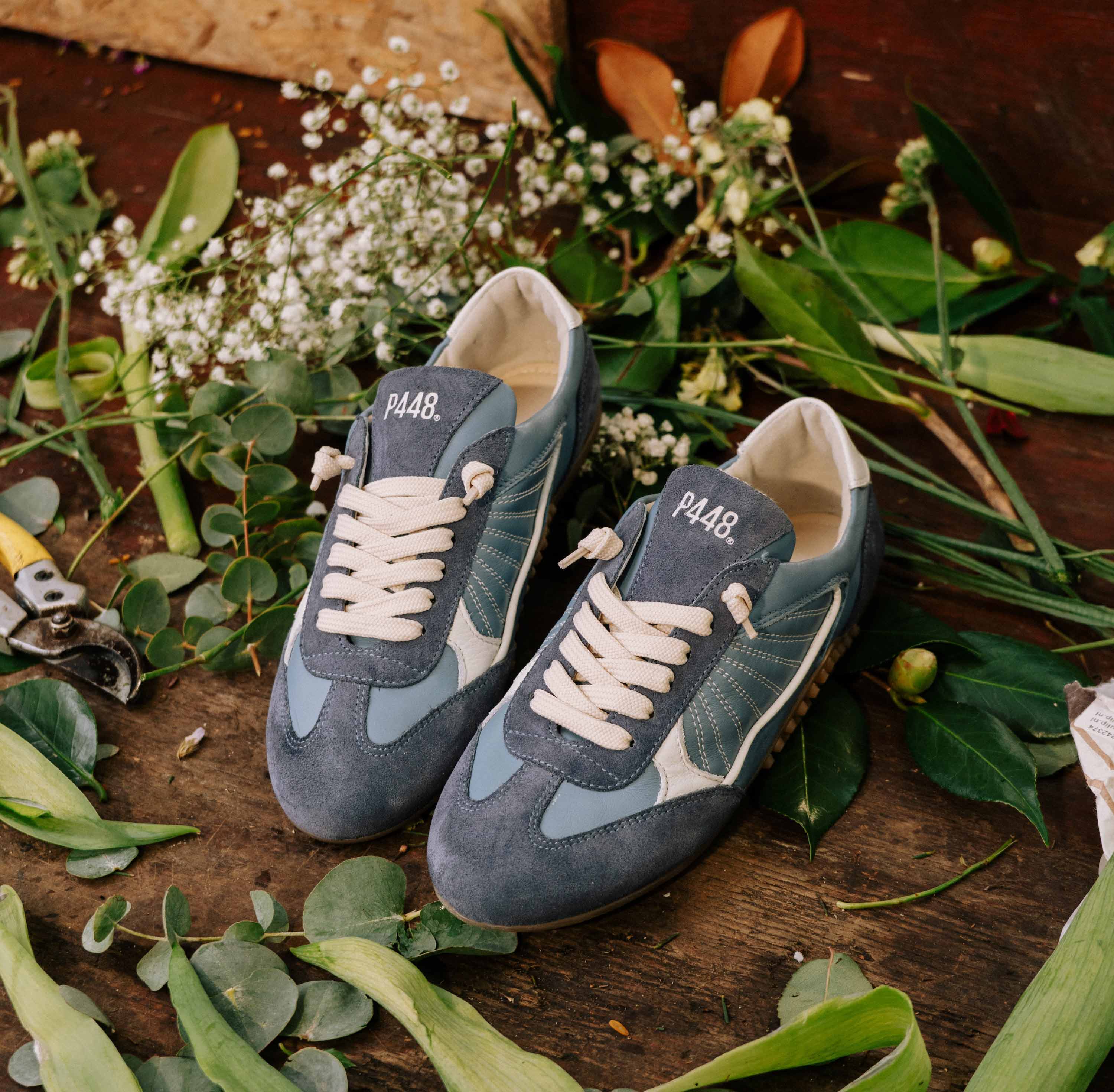 Top-down view of P448 blue leather and blue suede sneakers on a rustic wooden surface, surrounded by eucalyptus leaves and white flowers. Features cream laces, contrast stitching, and a white heel tab.