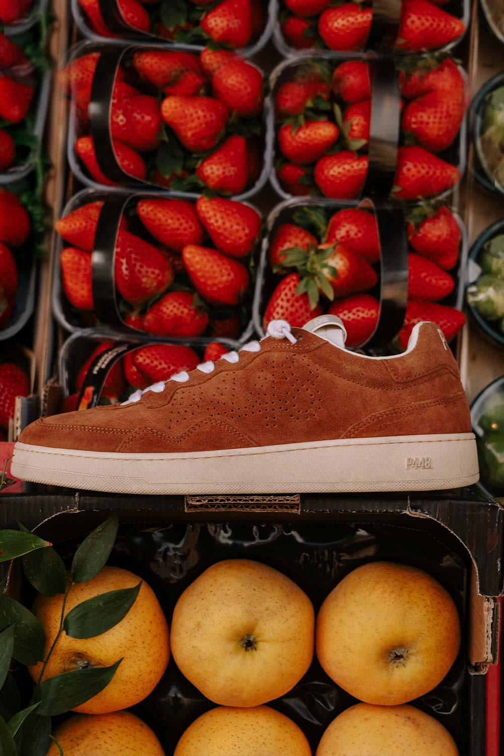 Red suede P448 low-top sneaker featuring white laces and a cream rubber sole with debossed branding, showcased in a vibrant market setting.