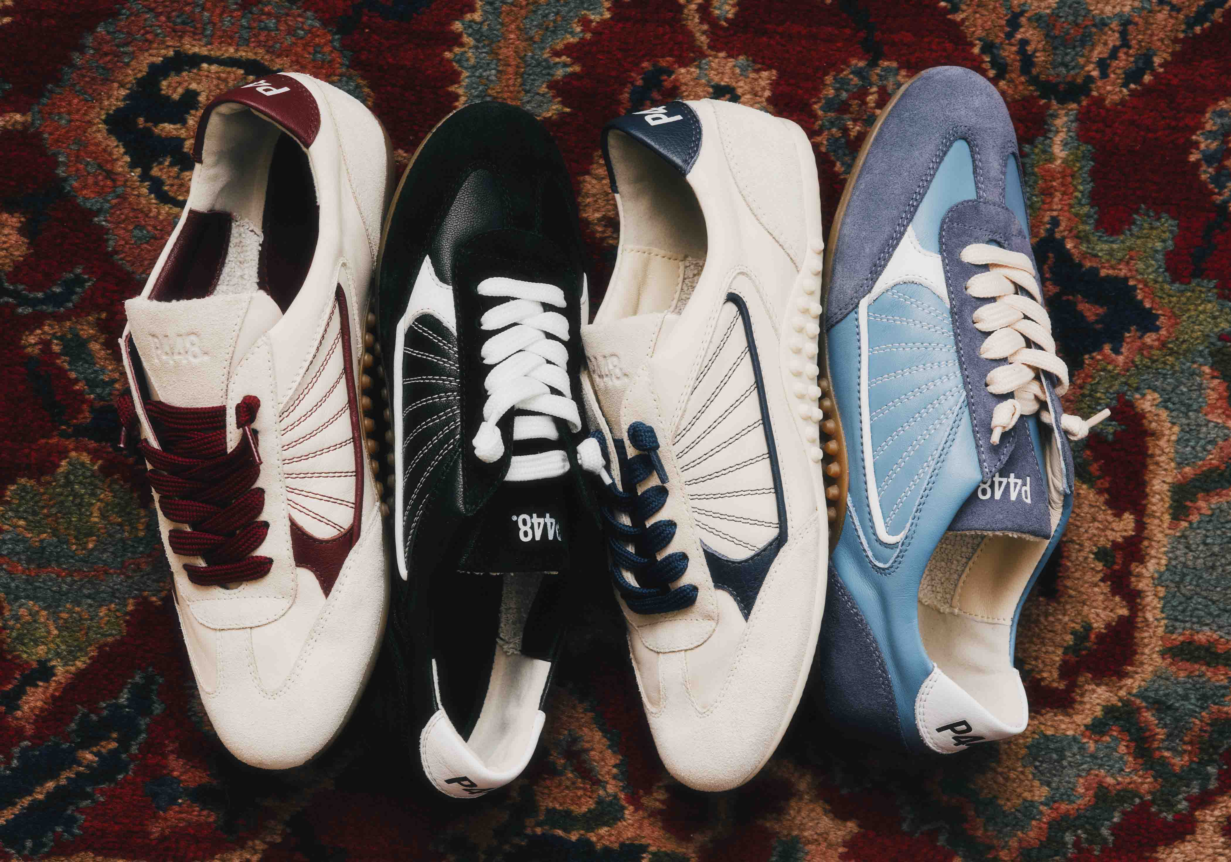 Four pairs of P448 Lisbon designer sneakers in burgundy, black, navy, and blue colorways on a vintage patterned rug.