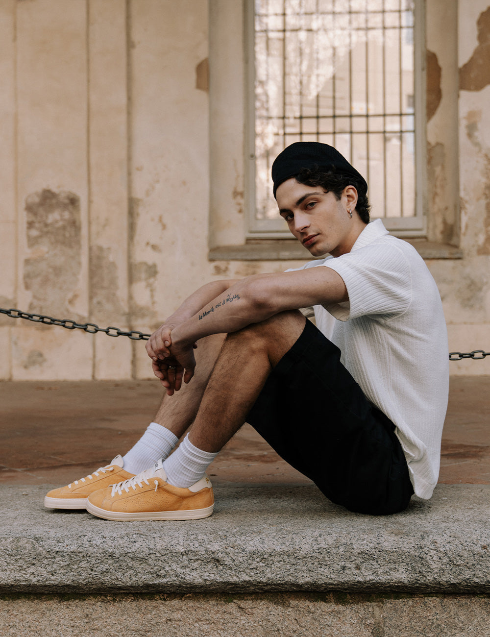 Stylish young man wearing bold mustard suede sneakers and a black beanie, sitting on a stone wall against an old building facade.