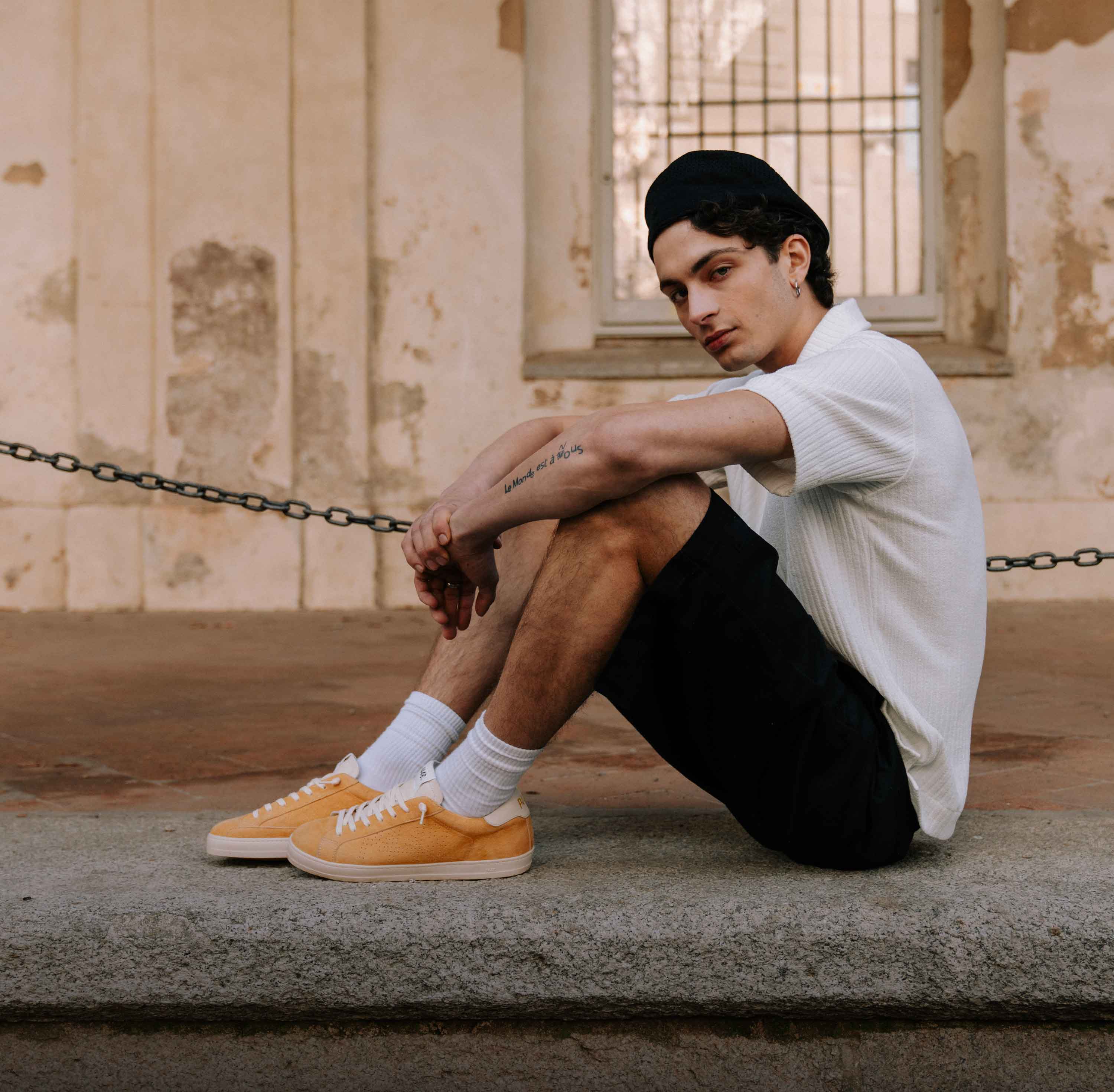 Stylish young man wearing bold mustard suede sneakers and a black beanie, sitting on a stone wall against an old building facade.