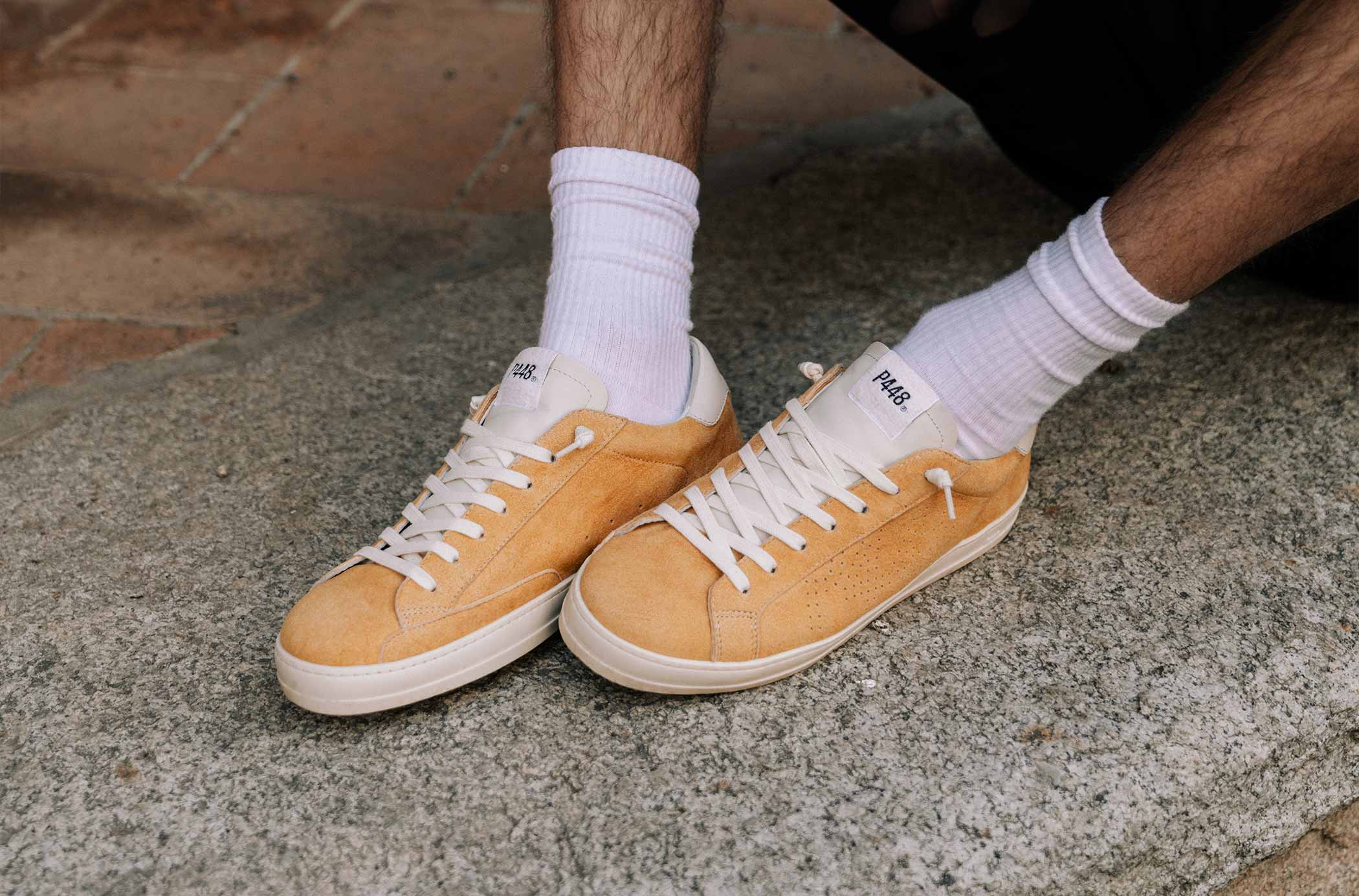 Close-up of a men wearing mustard yellow suede P448 sneakers with white laces and white ribbed socks, sitting on a stone surface.