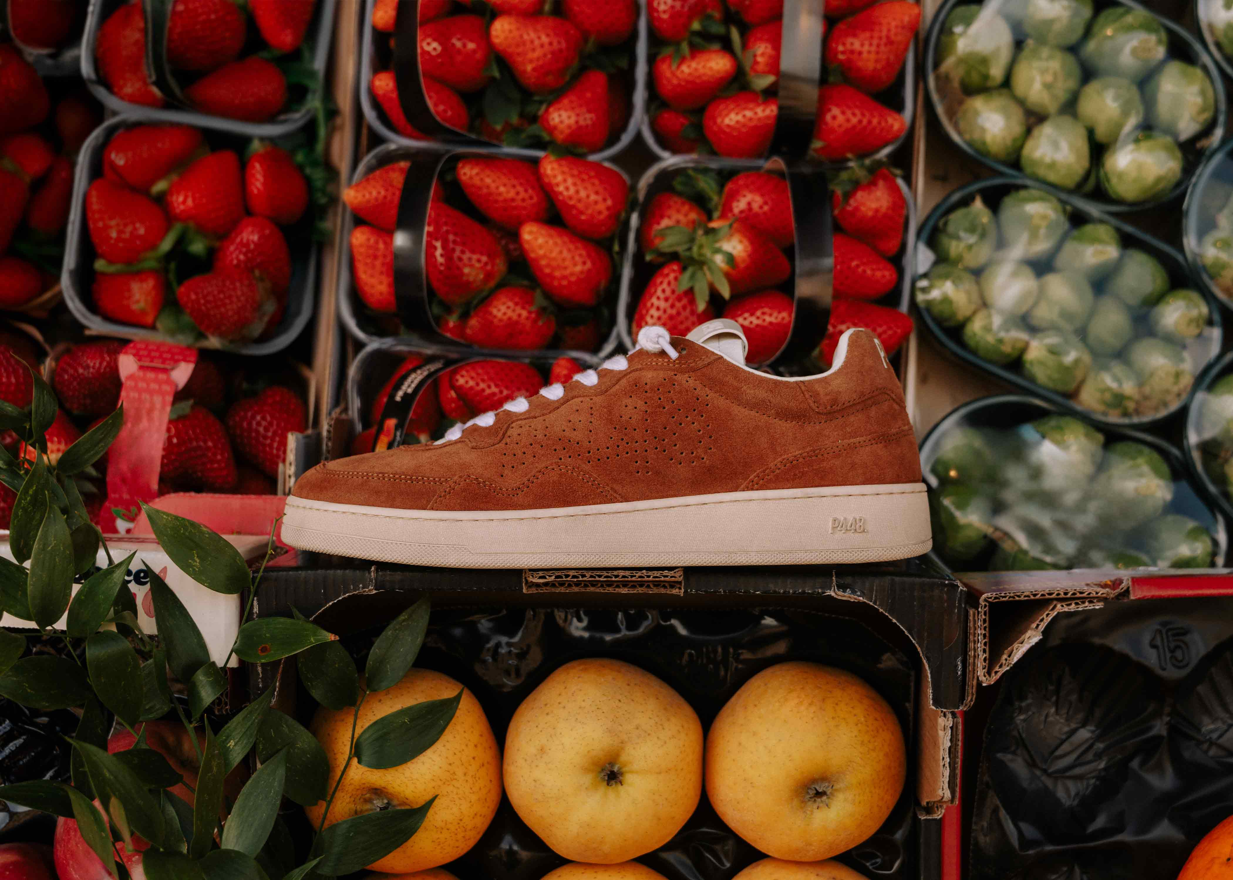 Red suede P448 low-top sneaker featuring white laces and a cream rubber sole with debossed branding, showcased in a vibrant market setting.